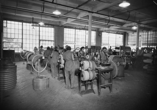 A group of women work at machines making tires. Men are in the background on the right working on other machines.