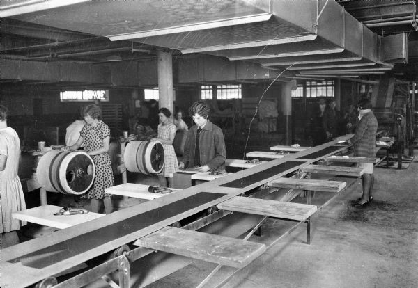 Women assemble tires at the Uniroyal plant. Men are also working in the background.