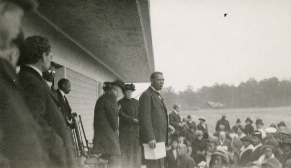 A man thought to be Booker T. Washington speaks to a crowd of people from the porch of a building.