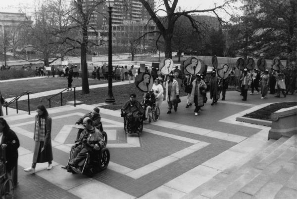 Members of Service Employees International Union (SEIU) Local 880 march across a plaza in protest. Some are carrying signs shaped like giant pennies. Disabled members in wheelchairs are present.