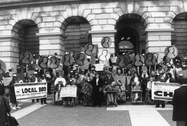 Group portrait in front of a large stone building with arches of members of the Service Employees International Union (SEIU) Local 880 holding giant pennies and banners. Several disabled activists in wheelchairs are present in the front row.