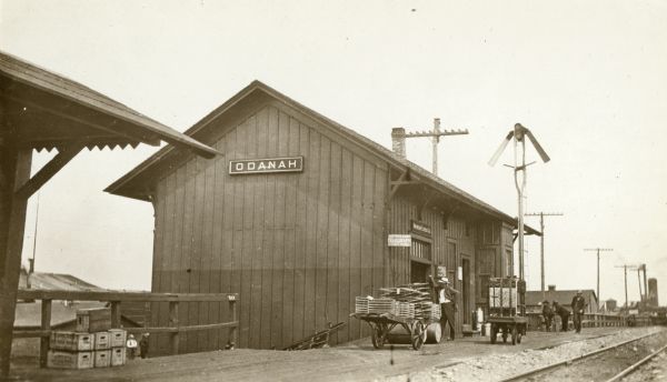 View over railroad tracks of railway depot. Men are standing around the station, and carts are parked on the station platform.