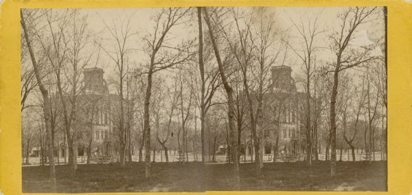 Stereograph of Madison City Hall at the corner of Mifflin Street and Wisconsin Avenue, as viewed through the trees of the Capitol Park. The three-story tall building was constructed in 1858. The building was sufficiently large that the third floor was entirely given over to a public auditorium and some of its offices could be rented to the state for extra income for the city. The old City Hall was not destroyed until 1954.