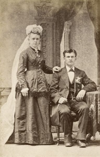 Studio wedding portrait in front of a painted backdrop of Gustav Willms (c. 1854-1917) and Kunigunde (Dorn) Willms (1857-1941) in their wedding outfits. Kunigunde wears a veil. The couple were married on April 17, 1880 in Milwaukee, Wisconsin.