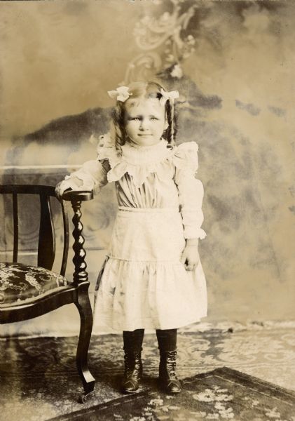 Full-length studio portrait in front of a painted backdrop of Lydia Willms (b. 1896). She is resting her hand on a chair, and is wearing a dress, ribbons and ringlets in her hair, and high button boots.

She was the daughter of Gustav Willms (c.1854-1917) and Kunigunde (Dorn) Willms (1857-1941).  Gustav ran a cigar factory at the back of their house.
