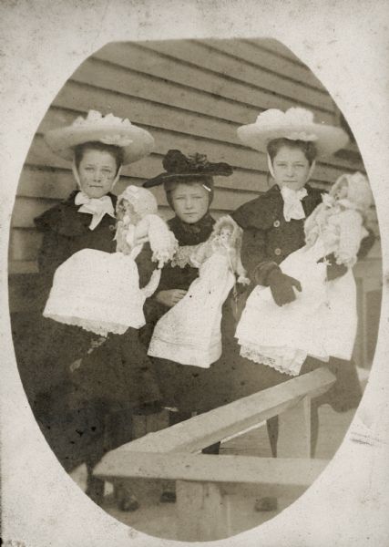 Lydia Willms (b. 1896), center, with her twin sisters Emma (1893-1974) and Marge (1893-1973) holding their dolls posing standing outside a building, probably their house in Milwaukee, Wisconsin. They are wearing fancy hats with flower decorations. The dolls are dressed in bonnets and long, white dresses.

They were the daughters of Gustav Willms (c. 1854-1917) and Kunigunde (Dorn) Willms (1857-1941). Gustav ran a cigar factory at the back of the Willms home on Muskego Avenue, Milwaukee.