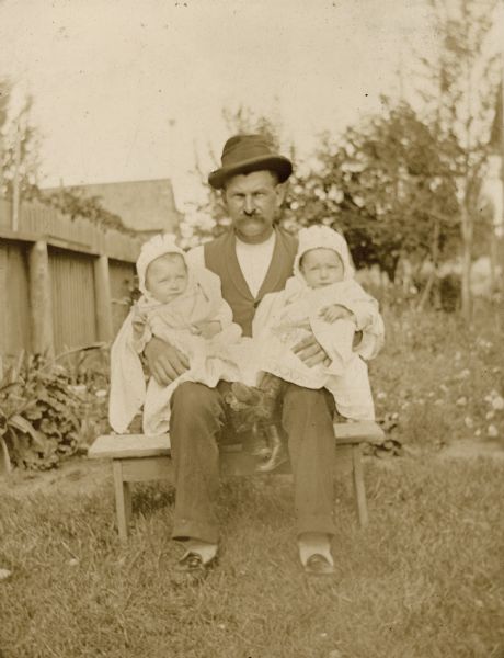 Gustav Willms (c. 1854-1917) of Milwaukee, Wisconsin, seated outdoors holding twin daughters Marge and Emma (b. December 27, 1893), who are wearing dresses, bonnets and high button shoes. Gustav owned a cigar factory.