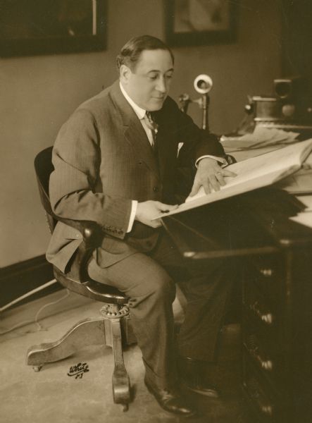Henry B. Harris is seated at a desk reading a large book or ledger. He is wearing a suit, and there is a telephone at his elbow.