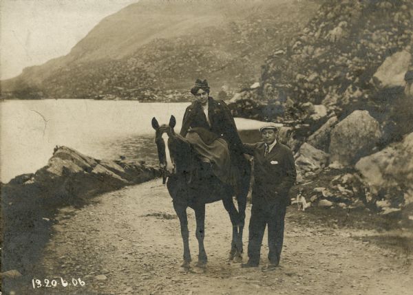 Renee and Henry Harris pose on a rocky beach in Killarney. She is seated sidesaddle on a horse and Henry stands at her side. A small dog sits behind them in the road.
