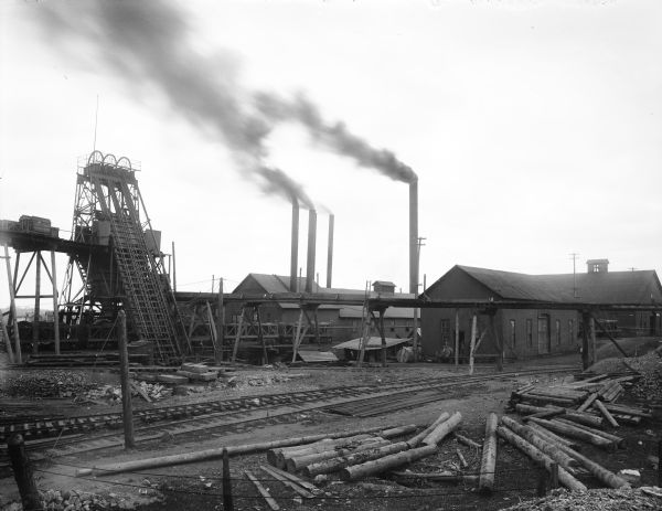 Exterior view of the Ashland Mine. Railroad tracks and piles of lumber are in the foreground, and chimneys spew smoke from buildings in the background.