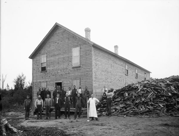 A group of people pose in front of a boarding house. One man on the right is holding up a small child. There is a large pile of lumber or fuelwood next to the building. A man wearing an apron holds a long horn next to him.