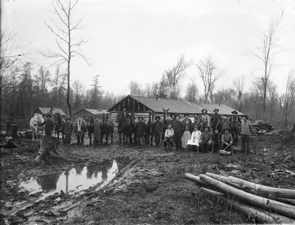 A group of people, presumably loggers and camp employees, pose with horses at a logging camp. A muddy puddle is in the foreground, and behind the group are log buildings.