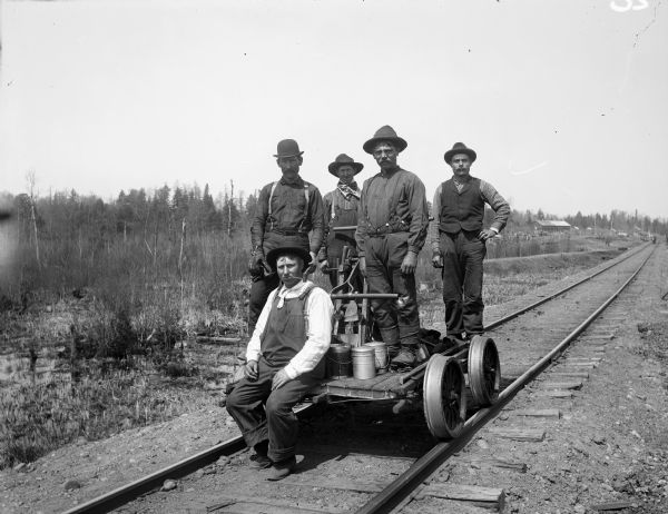 Five men known as the Railroad Section Gang pose together on a handcar on railroad tracks. They traveled on handcars and repaired tracks. Standing second from left is John Barto, and third from left is Frank Organist. The other men are unidentified. There is a large building behind the group on the right in the far background.