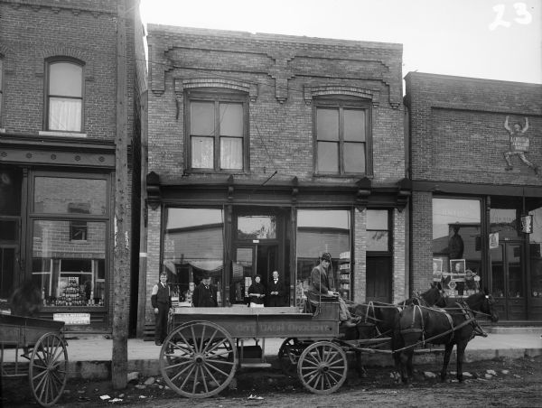 View from street of man seated on a horse-drawn wagon on Silver Street, which is parked in front of (from left to right) Joseph Rainieri Meat Market, Egan and Reardon Grocery, and Whitman's Drug Store. Several people pose in front of the grocery store.