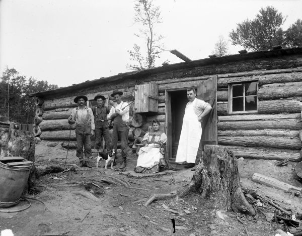 A woman (seated) and four men pose in front of a log building with a tree stump in the foreground. One man is holding a rifle, and is also holding a dog on a leash standing at his feet.
