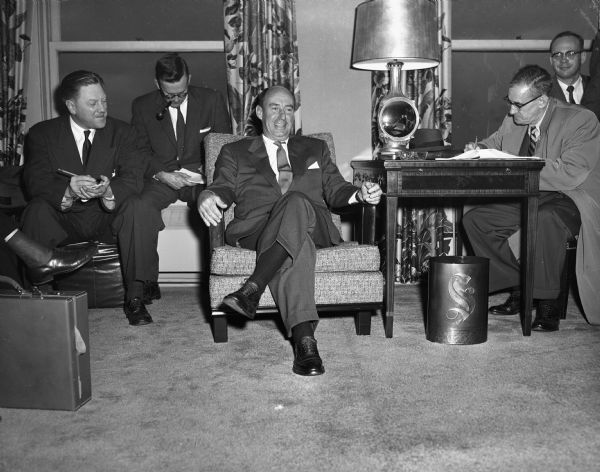 Adlai Stevenson is seated in a room with several other men.
