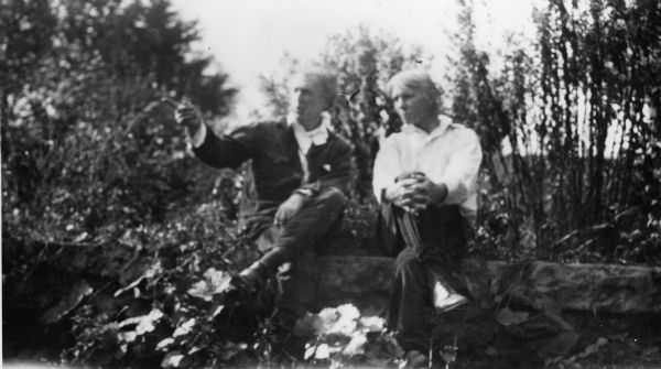 Frank Lloyd Wright and Carl Sandburg sit together on a stone wall outdoors surrounded by plants and trees.