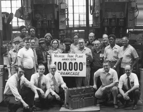 A group of male and female workers are posing around a sign and a crankcase sitting on the factory floor. The sign reads "Melrose Park Plant / March, 1974 / 100,000th Crankcase." Five men wearing ties are kneeling in front.
