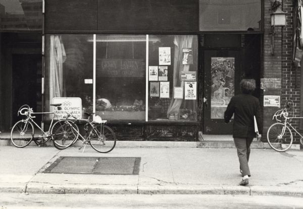 A man walks towards the Green Lantern Eating Cooperative. There are posters in the large front window and several bicycles are parked in front of the co-op.
