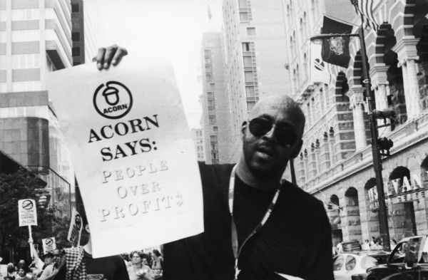 A man wearing sunglasses holds up a sign that reads "ACORN Says: People Over Profits" at a rally.