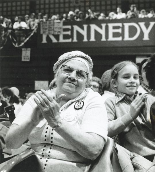 An elderly woman wearing an ACORN button sits next to a little girl at the Democratic delegation. They are clapping and there is a "Kennedy" banner behind them. The name "Willma Lee..." is written on the border of the photograph.
