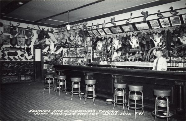View of the bar at John Kinziger and son's Hunters and Fishermans Tavern. A man, presumably John Kinziger, stands behind the bar. The walls are covered with various taxidermied animals. There are spittoons on the floor along the front of the bar. Caption reads: "Hunters and Fishermans Tavern John Kinziger and Son — Lena — Wis."