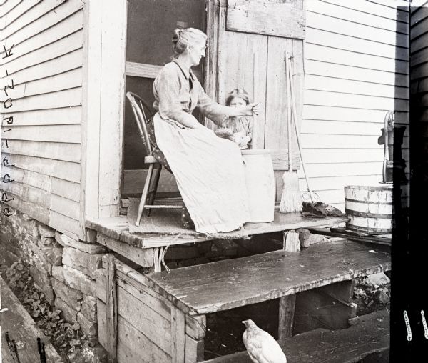 Woman using a dasher churn to make butter. She is seated on the stoop of a house and a young girl watches behind her.