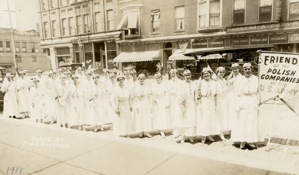 Group of women gathered at 2nd (now 7th St.) and Mitchell wearing light-colored dresses and hats. Each woman holds a U.S. flag. There is a sign to the right reading "Friends of the Polish companies in the U.S. Army." Across the street behind the women are several businesses, including a buffet, A.J. Graettinger Liquors, T. Dembinski's (?), Forecki & Przyby(?), and a chop suey restaurant.