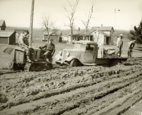 A man with a tractor attempts to pull a truck out of deep mud. Two men stand on the bed of the truck, and another man sits on the front of the tractor. These poor road conditions occurred on Highway 80 near Elroy.