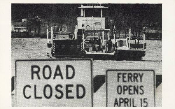 The Merrimac ferry "Colsac II" is seen on the Wisconsin River behind two signs. The signs read, "Road Closed" and "Ferry Opens April 15".