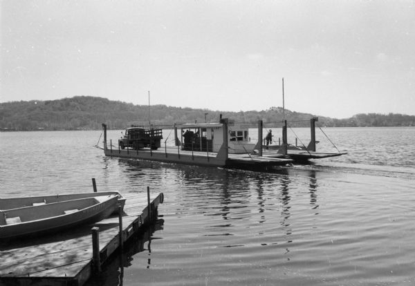 The Merrimac Ferry is on the river with a pick-up truck aboard. A rowboat rests on a pier in the foreground. A man wearing a hat rests his foot on the railing. In the foreground two rowboats rest on a pier.