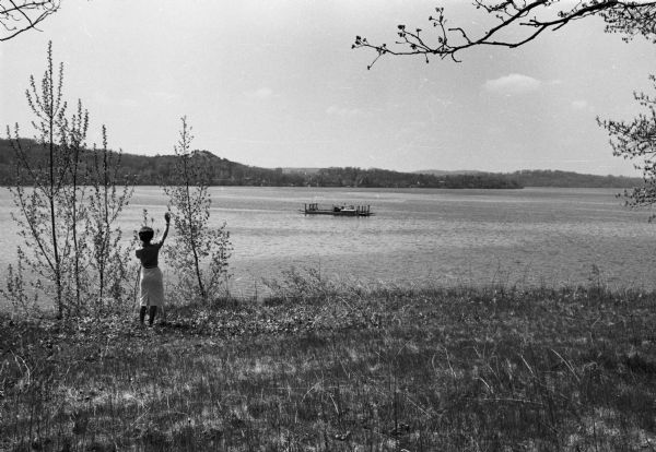 View from shoreline of a woman standing on a riverbank waving at the Merrimac ferry.