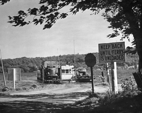 View from road of the Merrimac ferry approaching shore with several automobiles and a large truck aboard. In the foreground is a sign that reads: "Keep Back Until Ferry Is Unloaded".