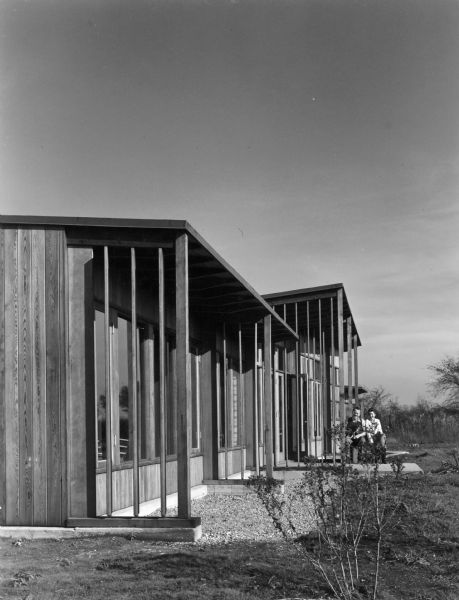 A man, holding a cat, is sitting next to a woman outside of a house which has a long row of tall windows. This Keck and Keck design was built for Hugh Duncan in Flossmoor, Illinois. Project #268.