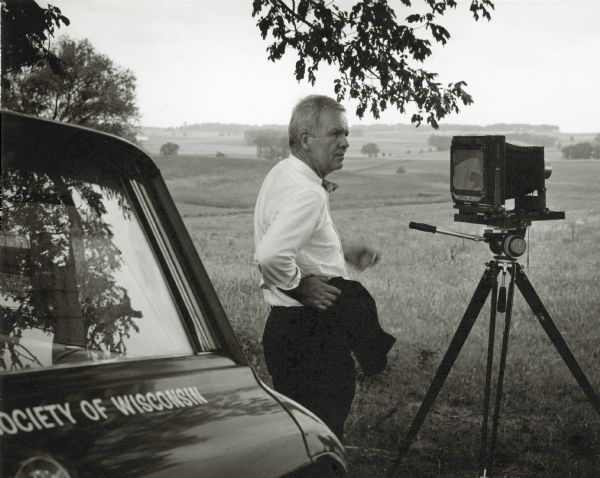 A candid portrait of Paul Vanderbilt in the field with his large format camera set up. He is standing next to a State Historical Society of Wisconsin vehicle.