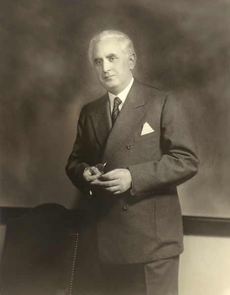 Three-quarter length portrait of Leo Crowley, chairman of the Federal Deposit Insurance Corporation of the United States. He is standing, holding his eyeglasses in his hands.