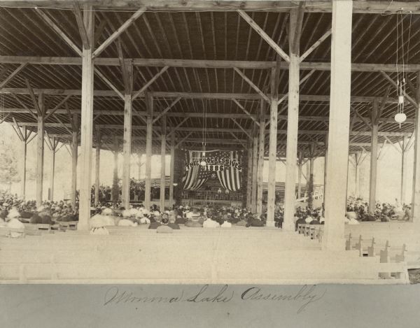 A view inside the Monona Lake Assembly Pavilion showing people gathered listening to speakers. Behind the speakers, a large banner is displayed. It is decorated as an American flag and reads, "Progressives, 1886".