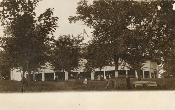An exterior view of people sitting and standing outside of the Monona Lake Assembly Auditorium, which is partially obscured by trees.  Designed by Morrison H. Vail of Chicago, built in 1900 by John H. Starck Co., John H. Findorff, vice-president.  Torn down in 1943.