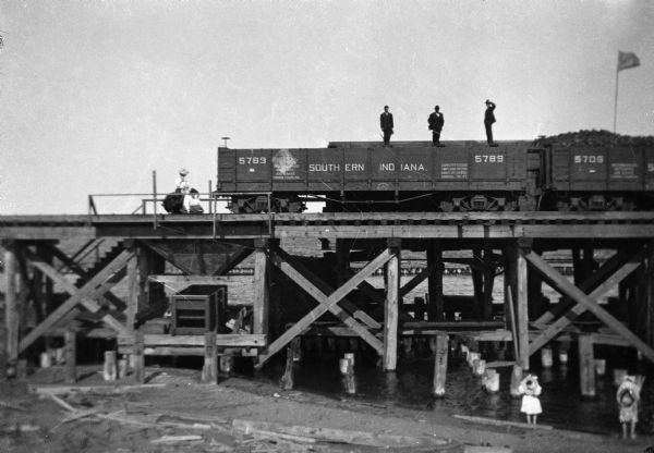 View from below of several freight cars on a railroad bridge over a river, with three men standing on top of a railroad car. A woman with a child is standing on the bridge on the left. Two children are standing below the bridge on the shoreline.