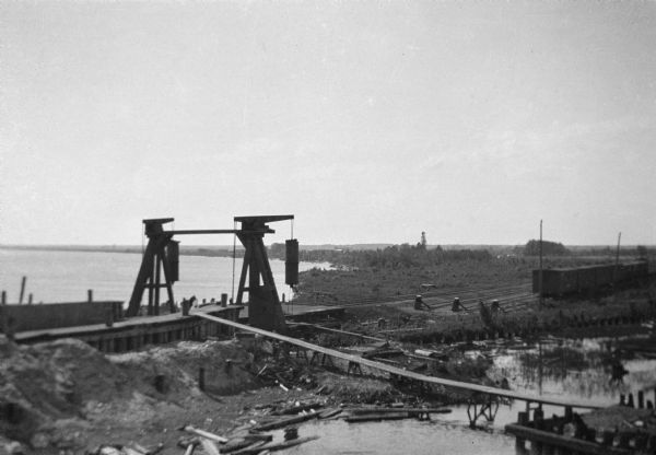 Elevated view of an early phase of construction of a railroad bridge. In the background is a body of water, and in the foreground are wooden walkways over more water. Construction debris is scattered around the site. Railroad cars are on railroad tracks on the right.