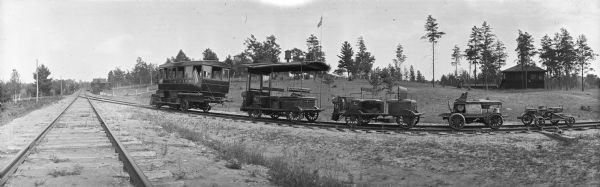 Panoramic view of a variety of small railroad cars on a set of curved railroad tracks that are merging into railroad tracks that stretch to the horizon. In the background is a flag on a flagpole, a water tower, and a building.