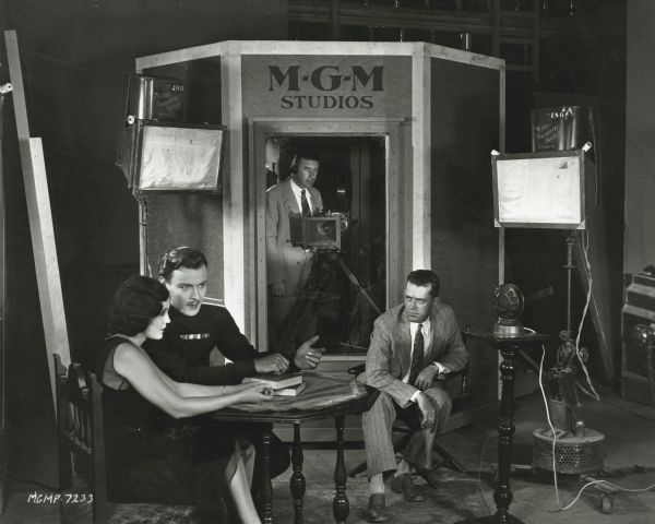 A man operates a Mitchell camera from inside an MGM sound booth as Raquel Torres and Nils Asther perform seated in front of lights and a microphone. Director W.S. Van Dyke sits next to the microphone and watches intently.