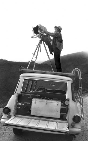 Ansel Adams inserts the film holder into his 8 x 10 camera. He is photographing from a platform mounted on the top of his International Travelall truck.