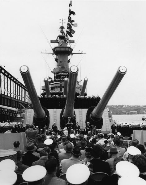 Ceremony beneath the guns on board the U.S.S. <i>Wisconsin</i>. A number of spectators and sailors are seated in the foreground, and uniformed sailors line both sides of the ship, as well as the gun turrets.