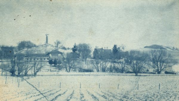 Cyanotype of the southeast elevation of Hillside Home School in winter. The Romeo and Juliet windmill is on a hill in the background. A field and fence are in the foreground.