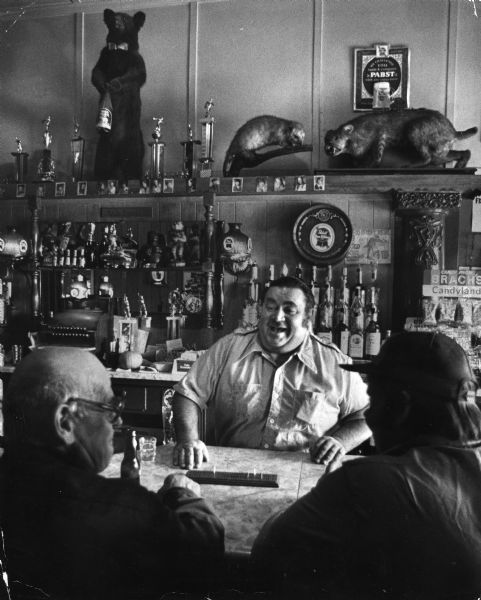 Bartender Rod Ballmer (center), Paul Bernsdorf (left), and Jeffrey Mondeik at Ballmer's Old Time Tavern. There is a cribbage board on the bar and the wall behind the bar is covered with bottles, knickknacks and taxidermied animals.