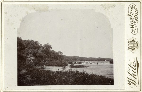View of the Sugar River below a dam. A man stands in the water fishing. Two others stand on the bank at left and one is seated on the dam. Three sticks are tied together as a tripod on the hill at left.