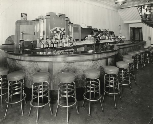 Interior view of an art deco-style bar lined with stools. There is a radio behind the bar along a mirrored wall where bottles of liquor are displayed around vases of flowers. The mens' room is at the end of the bar on the right if you need it. Stairs to an upper level can be seen in the upper right corner.