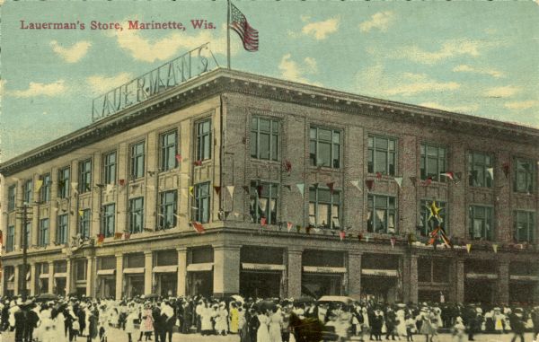 Exterior view across intersection toward the Lauerman's Store on a street corner. There are pennant strings hanging on upper floors of the building, and a U.S. flag is flying on the roof. A large group of people are congregating in the street. Caption reads: "Lauerman's Store, Marinette, Wis."