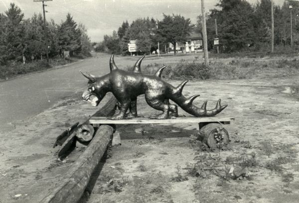 Hodag standing on a board balanced on logs near a road. There is a gasoline filling station in the background.